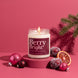 Berry Bright soy candle in an 8 oz glass jar with a plum label, lit on a pink pedestal with sugared plums, a halved blood orange, pink ornaments, and a pine sprig against a raspberry pink background
