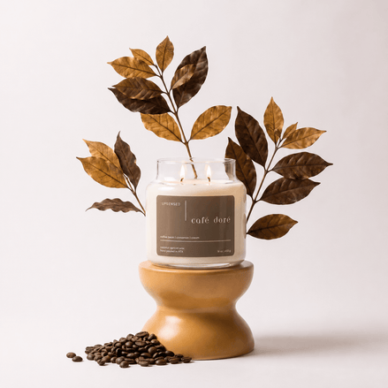 Upsensed Café Doré candle burning on a mustard-colored ceramic pedestal, styled with tall dried gilded leaf branches and scattered coffee beans at the base.
