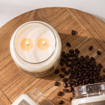 Overhead view of a burning Upsensed Café Doré candle on a round wooden board surrounded by scattered coffee beans, with additional candle jars at the edges of the frame.