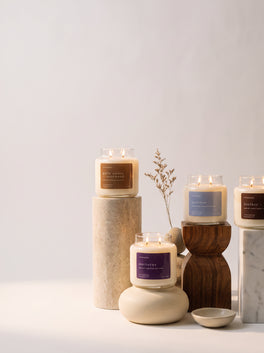 Four lit Upsensed candles — Palo Santo + Rosewood, Nocturne, Undertow, and Leather + Tabac — displayed at staggered heights on stone, wood, and marble pedestals with a dried botanical sprig.