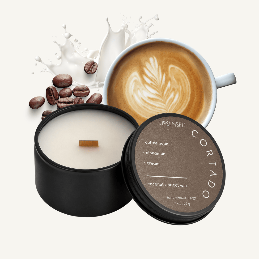 Upsensed Cortado 2 oz mini candle in a matte black tin with warm brown lid, surrounded by roasted coffee beans, a latte art cappuccino, and a splash of cream