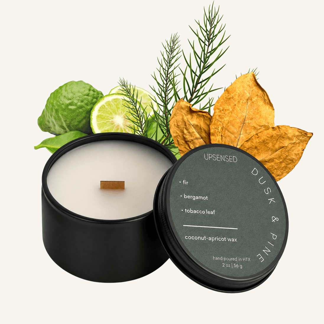 Upsensed Dusk & Pine 2 oz mini candle in a matte black tin with slate green lid, surrounded by fir needle sprigs, fresh bergamot, and dried tobacco leaves
