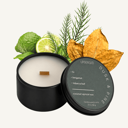 Upsensed Dusk & Pine 2 oz mini candle in a matte black tin with slate green lid, surrounded by fir needle sprigs, fresh bergamot, and dried tobacco leaves