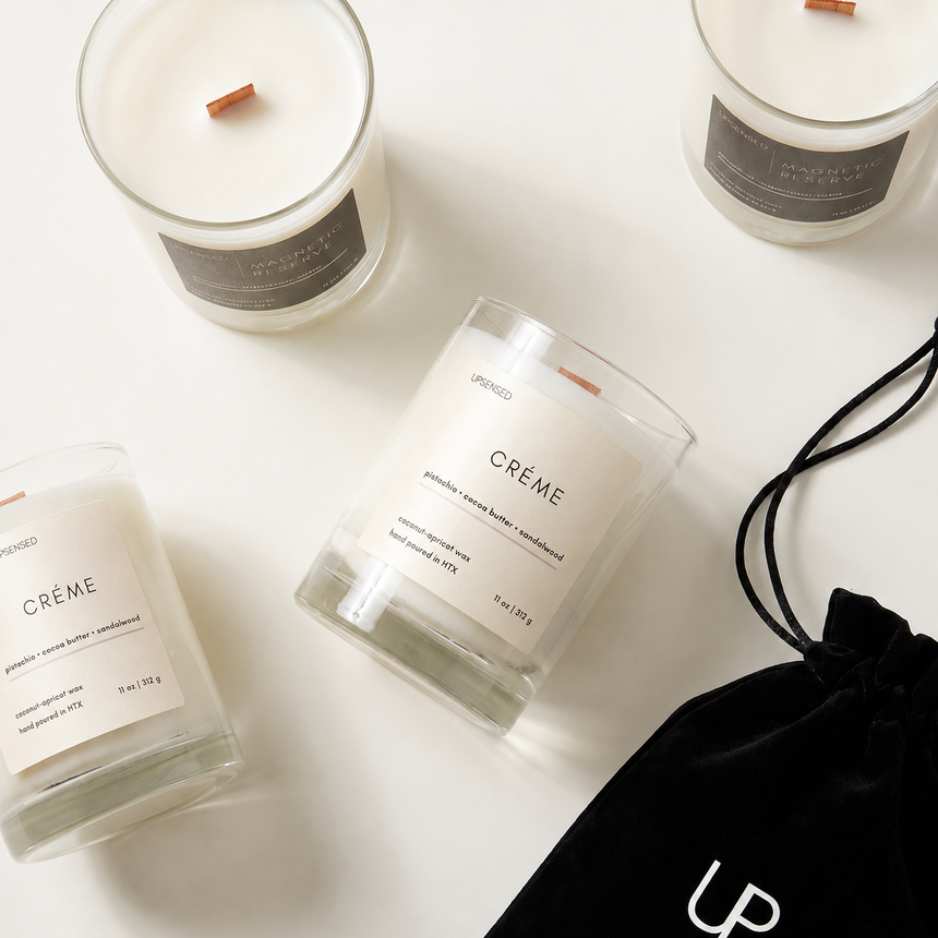 Upsensed full-size candles in glass vessels — Crème (pistachio, cocoa butter, sandalwood) and Magnetic Reserve — hand poured in Houston with coconut-apricot wax and wood wicks, shown with black velvet Upsensed pouch