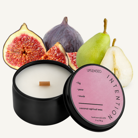 Upsensed Intention 2 oz mini candle in a matte black tin with blush pink lid, surrounded by halved fresh figs and whole green pears
