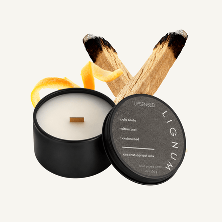 Upsensed Lignum 2 oz mini candle in a matte black tin with dark charcoal lid, surrounded by burned palo santo sticks and a curled citrus peel