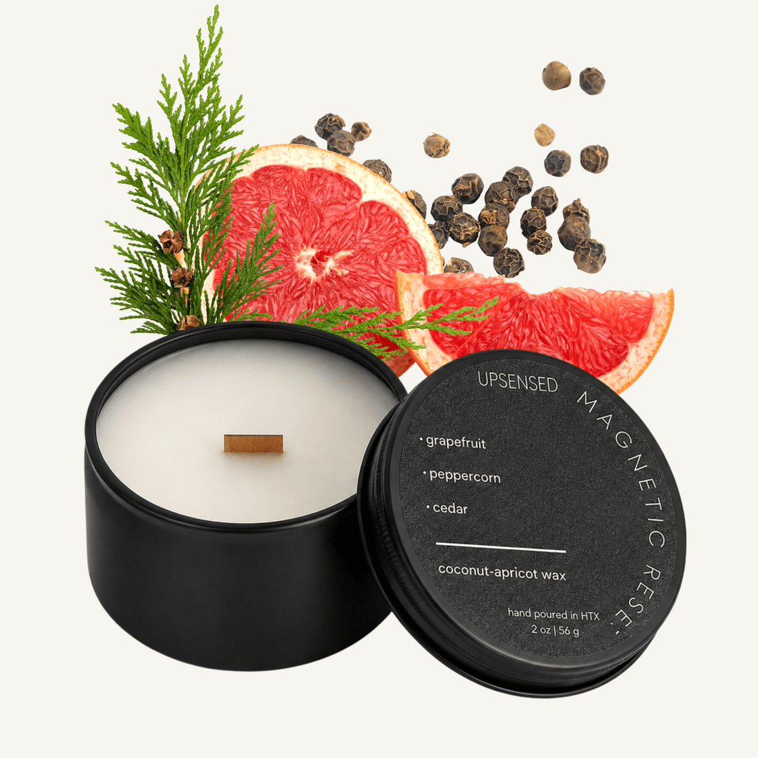 Upsensed Magnetic Reserve 2 oz mini candle in a matte black tin with dark lid, surrounded by fresh grapefruit slices, black peppercorns, and cedar sprigs