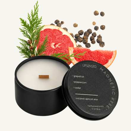 Upsensed Magnetic Reserve 2 oz mini candle in a matte black tin with dark lid, surrounded by fresh grapefruit slices, black peppercorns, and cedar sprigs