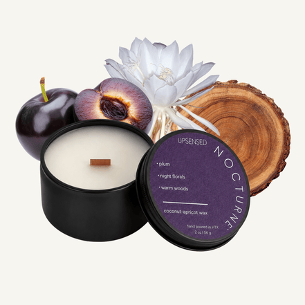 Upsensed Nocturne 2 oz mini candle in a matte black tin with deep purple lid, surrounded by whole and halved plums, a white Queen of the Night bloom, and a cross-section wood slice