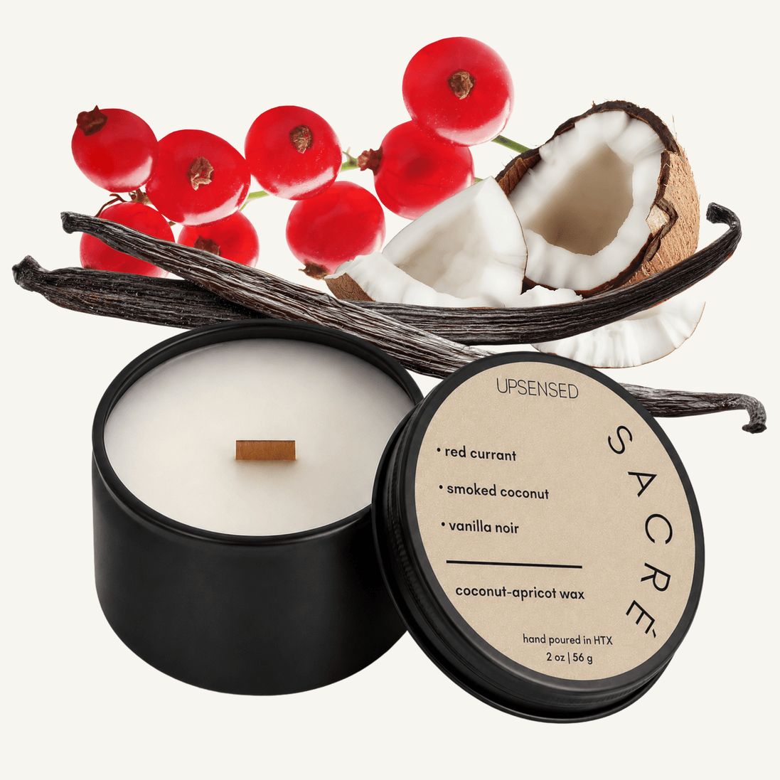 Upsensed Sacré 2 oz mini candle in a matte black tin with warm cream lid, surrounded by fresh red currants, a cracked coconut, and dark vanilla beans