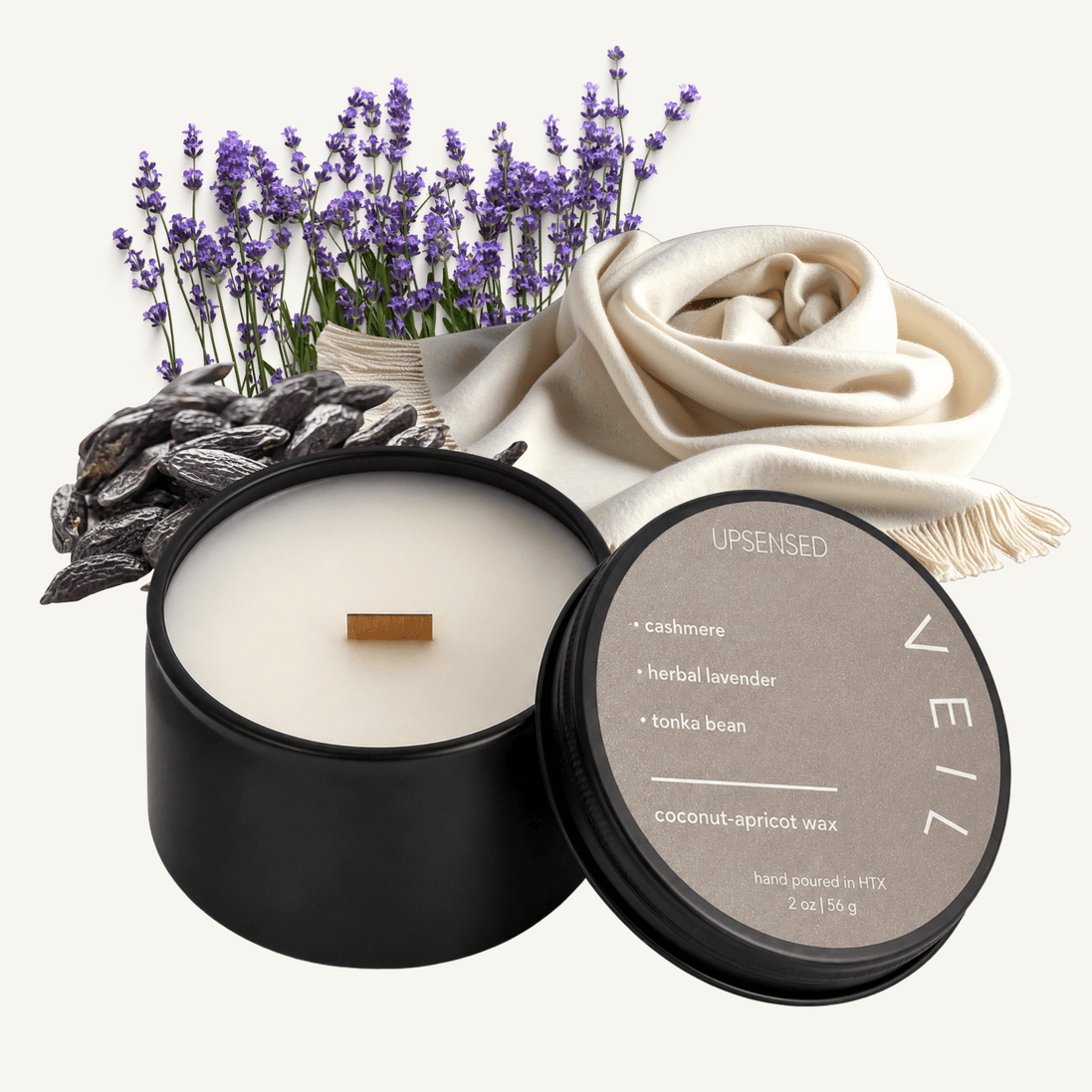 Upsensed Veil 2 oz mini candle in a matte black tin with warm taupe lid, surrounded by purple lavender sprigs, a cream cashmere scarf, and dried tonka beans