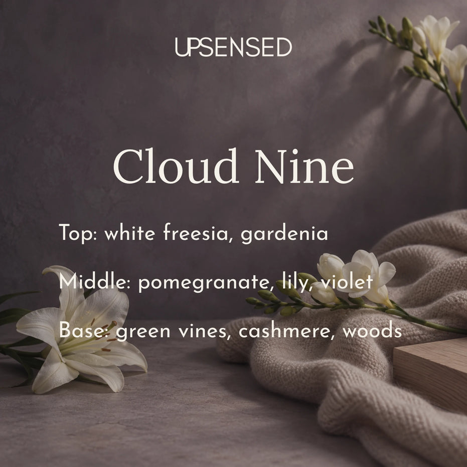 Cloud Nine soy candle fragrance notes: white freesia and gardenia on top, pomegranate, lily, and violet in the middle, and green vines, cashmere, and woods at the base, shown with white lilies, freesia, and a soft oatmeal cashmere throw against a dusky violet-gray backdrop.