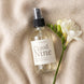 Cloud Nine 4 oz room and linen spray by Upsensed in a clear glass bottle with a soft cream label, lying on a draped oatmeal cashmere knit beside a single stem of white freesia, shot from above.