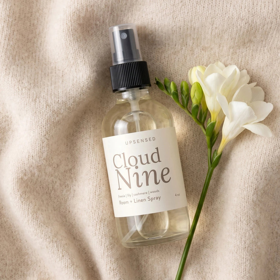 Cloud Nine 4 oz room and linen spray by Upsensed in a clear glass bottle with a soft cream label, lying on a draped oatmeal cashmere knit beside a single stem of white freesia, shot from above.