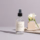 Cloud Nine 4 oz room and linen spray by Upsensed in a clear glass bottle with a black sprayer and a soft cream label listing freesia, lily, cashmere, and woods notes, beside a pale oak block, white freesia, and an oatmeal cashmere knit on a dove gray set.