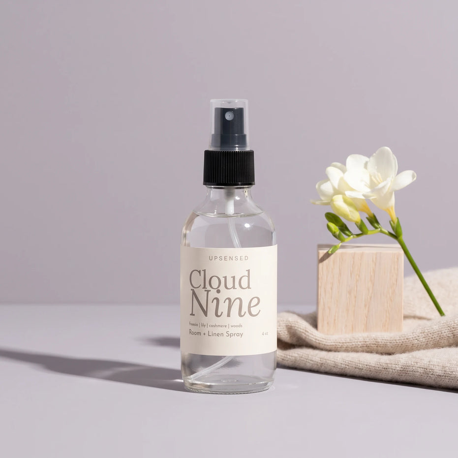 Cloud Nine 4 oz room and linen spray by Upsensed in a clear glass bottle with a black sprayer and a soft cream label listing freesia, lily, cashmere, and woods notes, beside a pale oak block, white freesia, and an oatmeal cashmere knit on a dove gray set.