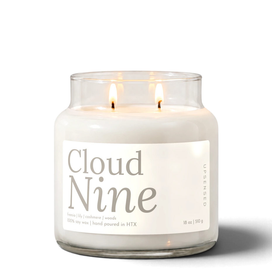 Cloud Nine 18 oz double-wick soy candle by Upsensed, both wicks lit, in a clear glass jar with a soft cream label in warm gray type listing freesia, lily, cashmere, and woods notes.
