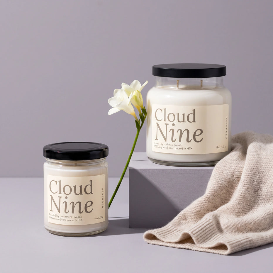 Cloud Nine soy candles by Upsensed in 8 oz and 18 oz sizes with black lids and soft cream labels listing freesia, lily, cashmere, and woods notes, the 18 oz on a dove gray riser beside a single stem of white freesia and a draped oatmeal cashmere knit.