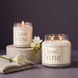 Cloud Nine 8 oz and 18 oz soy candles by Upsensed, lit, with soft cream labels in warm gray type, the 8 oz on a pale oak riser beside a folded oatmeal cashmere knit, a white lily, and a stem of white freesia against a dusky violet-gray backdrop.