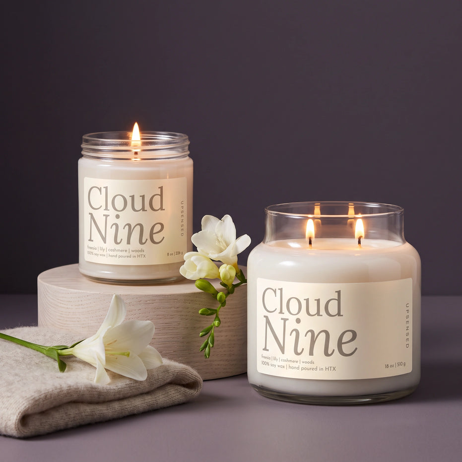 Cloud Nine 8 oz and 18 oz soy candles by Upsensed, lit, with soft cream labels in warm gray type, the 8 oz on a pale oak riser beside a folded oatmeal cashmere knit, a white lily, and a stem of white freesia against a dusky violet-gray backdrop.