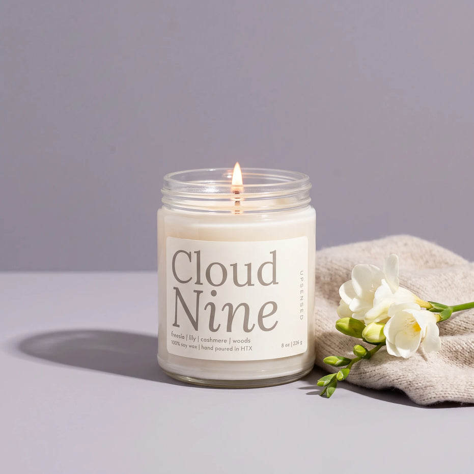 Lit Cloud Nine 8 oz soy candle in a clear glass jar beside white freesia and a cream knit throw on a soft lavender backdrop