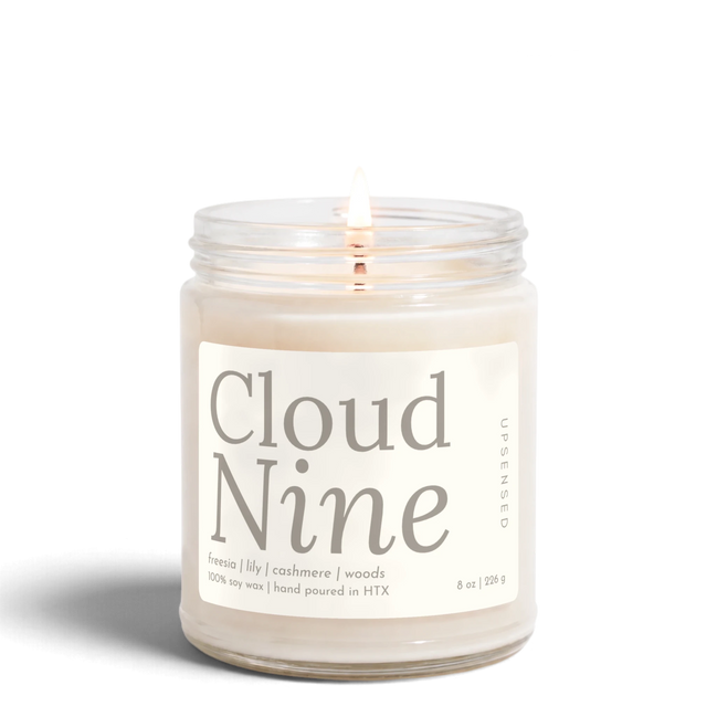 Cloud Nine 8 oz soy candle by Upsensed, lit, in a clear glass jar with a soft cream label in warm gray type listing freesia, lily, cashmere, and woods notes.