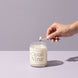 Hand lighting a Cloud Nine 8 oz soy candle with a match against a soft lavender backdrop
