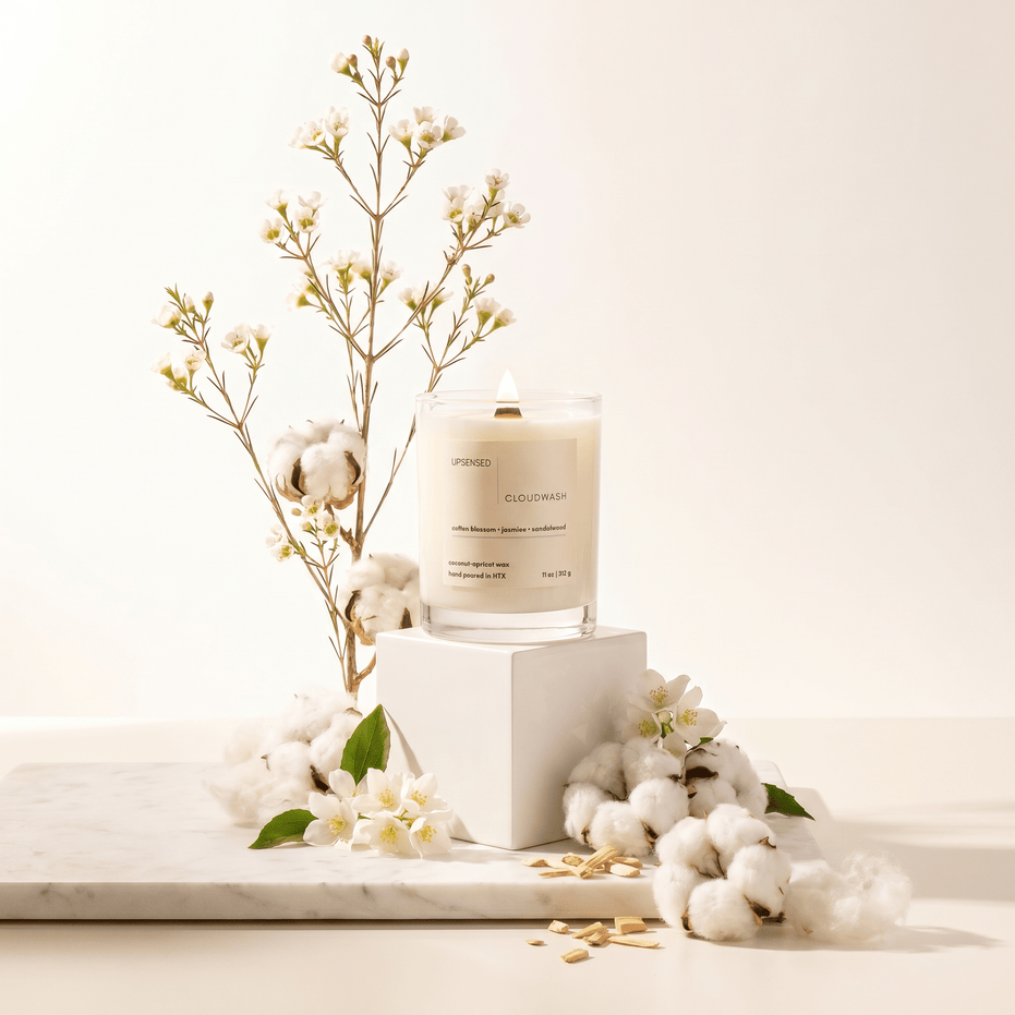 Upsensed Cloudwash candle on white pedestal surrounded by cotton bolls, jasmine flowers, and sandalwood chips on a marble surface