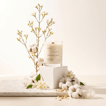 Upsensed Cloudwash candle on white pedestal surrounded by cotton bolls, jasmine flowers, and sandalwood chips on a marble surface