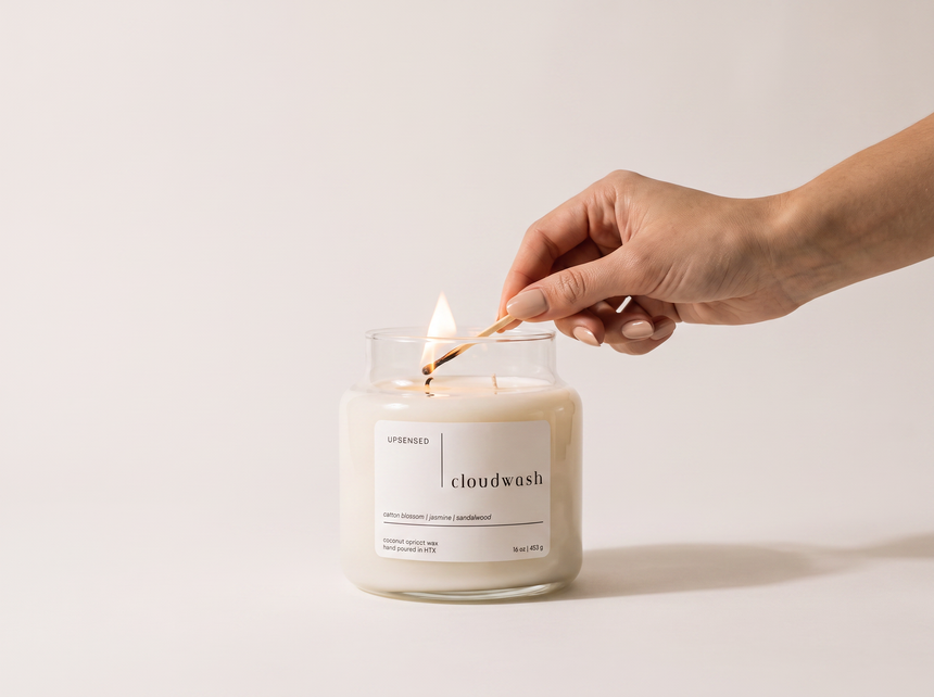 Hand lighting an Upsensed Cloudwash candle with a match against a soft neutral background, wax pooling around the wick.