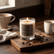 Upsensed Cortado gourmand candle burning on a dark wood board surrounded by scattered coffee beans beside a ceramic pour-over and cup