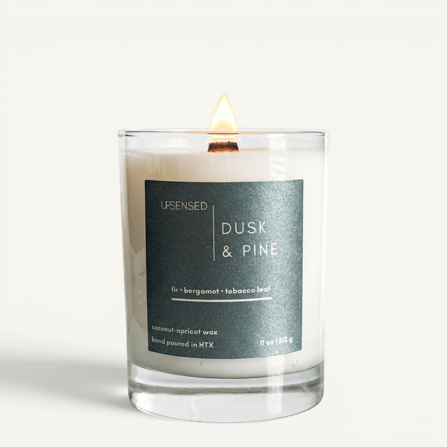 Upsensed Dusk & Pine candle lit on white background, showing deep teal label with fir, bergamot, and tobacco leaf notes