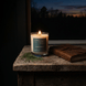 Upsensed Dusk & Pine woody candle burning on a rustic wood table beside a leather journal with a twilight forest visible through the window