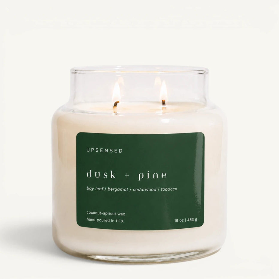 An Upsensed 'dusk + pine' scented candle in a clear glass jar with two lit wicks, featuring a dark green label on a clean white background.