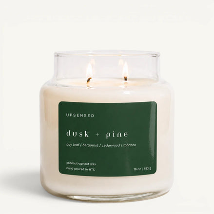 An Upsensed 'dusk + pine' scented candle in a clear glass jar with two lit wicks, featuring a dark green label on a clean white background.