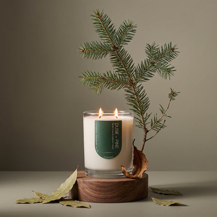 Lit Upsensed Dusk + Pine candle on a walnut pedestal with a fir branch, scattered bay leaves, and a curled dry leaf against a sage backdrop