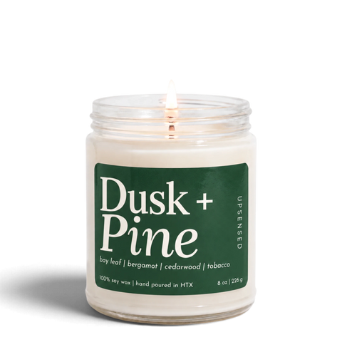 Upsensed Dusk + Pine 8 oz soy candle, lit, in a clear glass jar with a forest green label listing bay leaf, bergamot, cedarwood, and tobacco