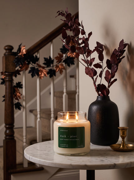 A lit candle in a clear glass jar sits on a round marble side table next to a matte black vase holding dark burgundy dried leaves, with a staircase and decorative garland in the background.
