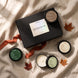 A curated fall discovery candle set from arranged on a beige linen fabric background. The set includes a black gift box with a white label, several open and closed black candle jars with green, brown, and cream labels, and scattered orange-brown autumn leaves with an orange ribbon.