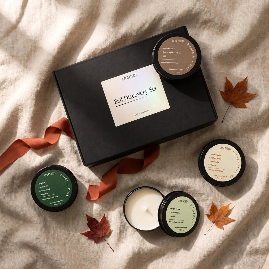 A curated fall discovery candle set from arranged on a beige linen fabric background. The set includes a black gift box with a white label, several open and closed black candle jars with green, brown, and cream labels, and scattered orange-brown autumn leaves with an orange ribbon.