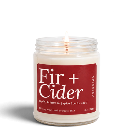 Upsensed Fir + Cider 8 oz soy candle, lit, in a clear glass jar with a cranberry red label listing apple, balsam fir, spice, and cedarwood