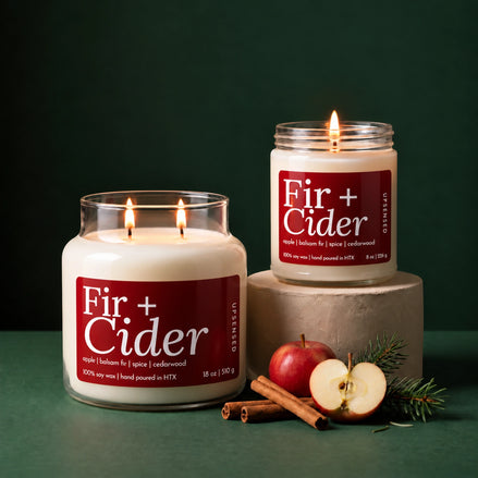 Two lit candles in clear glass jars with cream wax and dark red labels reading 'Fir + Cider' are styled on a dark green surface against a dark green background. One candle sits on a white pedestal, and the other is beside it.