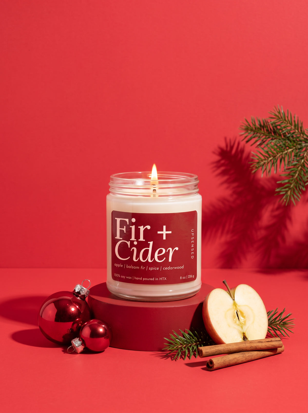 A lit candle in a clear glass jar sits on a red pedestal against a solid red background, styled with red ornaments, a sliced apple, cinnamon sticks, and evergreen branches. The label reads 'Fir + Cider'