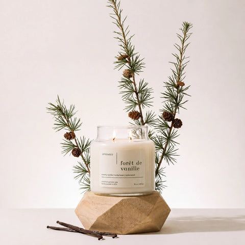 Upsensed Forêt de Vanille candle burning on a faceted wood pedestal, styled with pine branches, pinecones, and vanilla bean pods.