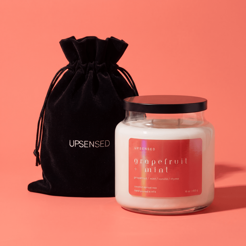 Upsensed Grapefruit + Mint candle with a black lid beside its black velvet UPSENSED drawstring packaging bag, on a coral background.