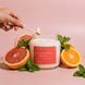 Hand lighting an Upsensed Grapefruit + Mint candle with a match, surrounded by halved grapefruits, a halved orange, and fresh mint sprigs against a pink background.