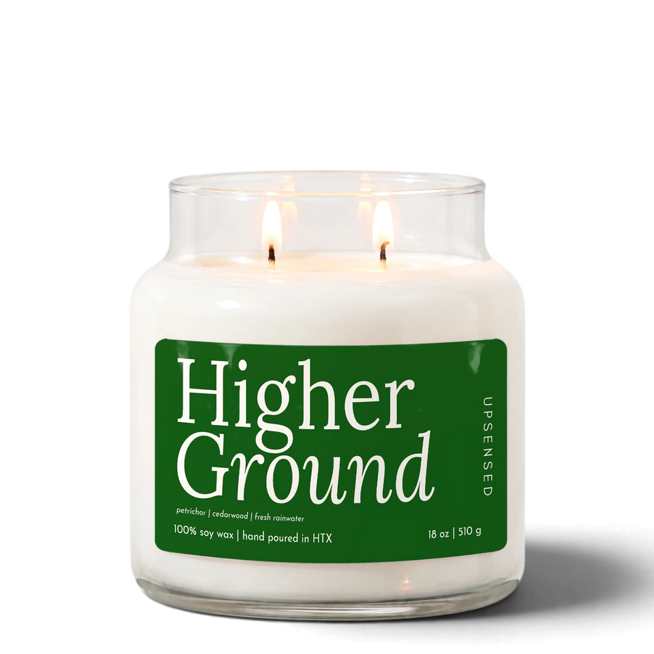 Higher Ground 18 oz double-wick soy candle by Upsensed, both wicks lit, in a clear glass jar with a forest green label listing petrichor, cedarwood, and fresh rainwater notes.