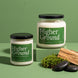 Higher Ground soy candles by Upsensed in 8 oz and 18 oz sizes with black lids and green labels listing petrichor, cedarwood, and fresh rainwater notes, styled with moss, cedarwood, and wet black stones on a sage green set.