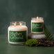 Higher Ground 18 oz and 8 oz soy candles by Upsensed, lit, with deep green labels, the 8 oz on a wet river stone beside moss, bark, and a fern frond against a dark forest green backdrop.