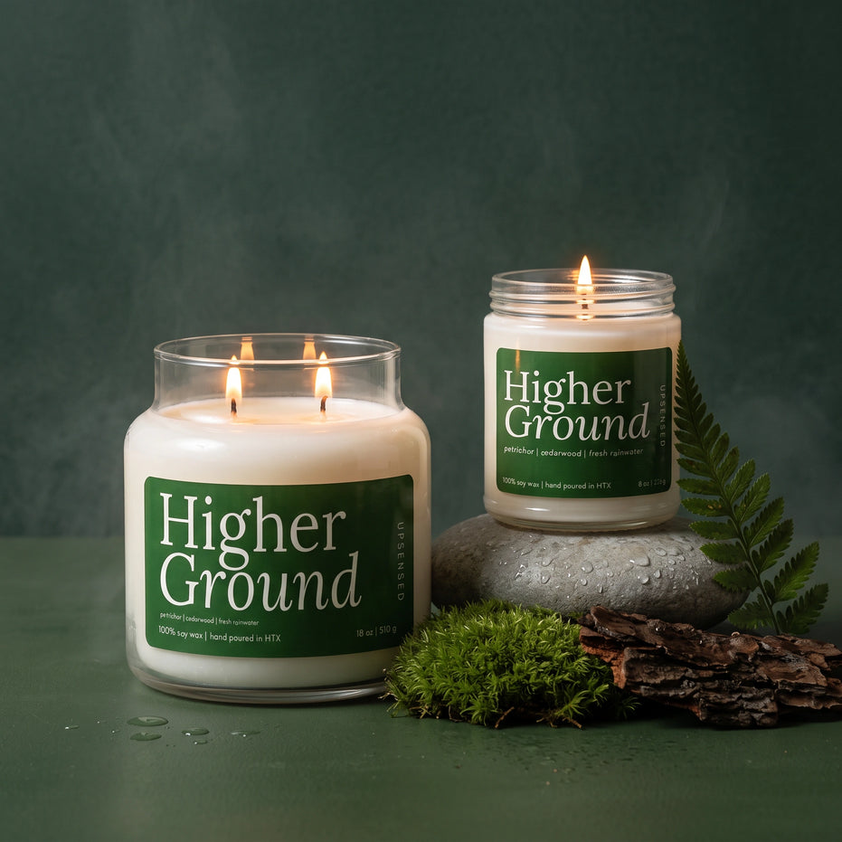 Higher Ground 18 oz and 8 oz soy candles by Upsensed, lit, with deep green labels, the 8 oz on a wet river stone beside moss, bark, and a fern frond against a dark forest green backdrop.